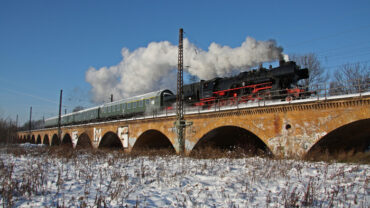 Das Bild zeigt den Museumszug mit Dampflok des Eisenbahnmuseums Leipzig auf einer Steinbrücke bei voller Fahrt.
