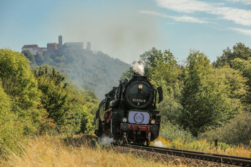 Sie sollten sehen: die Dampflokomotive des Eisenbahnmuseums Bayerischer Bahnhof zu Leipzig e. V. mit der Nummer 52 8154-8 in mitten vieler Bäume und Sträucher. Im Hintergrund trohnt die Wartburg auf einem Berg.