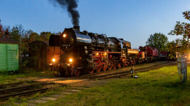 Sie sollten sehen: die Dampflokomotive 52 8154 bei Dämmerung mit ingeschaltetem Licht. Der Himmel ist tiefblau. die Lokomotive wird seitlich ein wenige angestrahlt. Im Hintergrund ist zu erkennen, dass sie den Eisenbahndrehkran (kurz EDK) Multitasker 1200 am Haken hat.