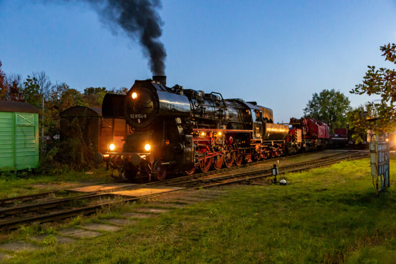 Sie sollten sehen: die Dampflokomotive 52 8154 bei Dämmerung mit ingeschaltetem Licht. Der Himmel ist tiefblau. die Lokomotive wird seitlich ein wenige angestrahlt. Im Hintergrund ist zu erkennen, dass sie den Eisenbahndrehkran (kurz EDK) Multitasker 1200 am Haken hat.