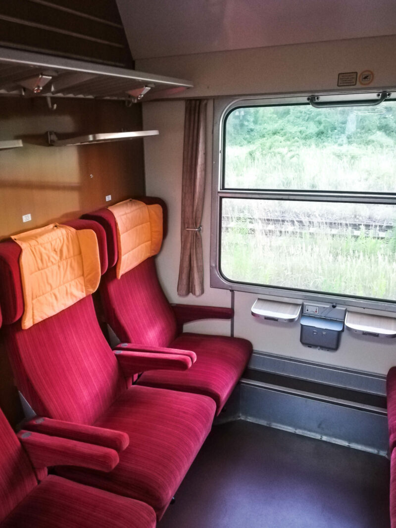 Blick in ein Wagenabteil mit sechs Plätzen. Bequeme Sessel mit roten Stoffbezügen und Orange Kopfkissen passen perfekt zur hellen Innenverkleidung.Städteexpresswagen Typ Y/B 70