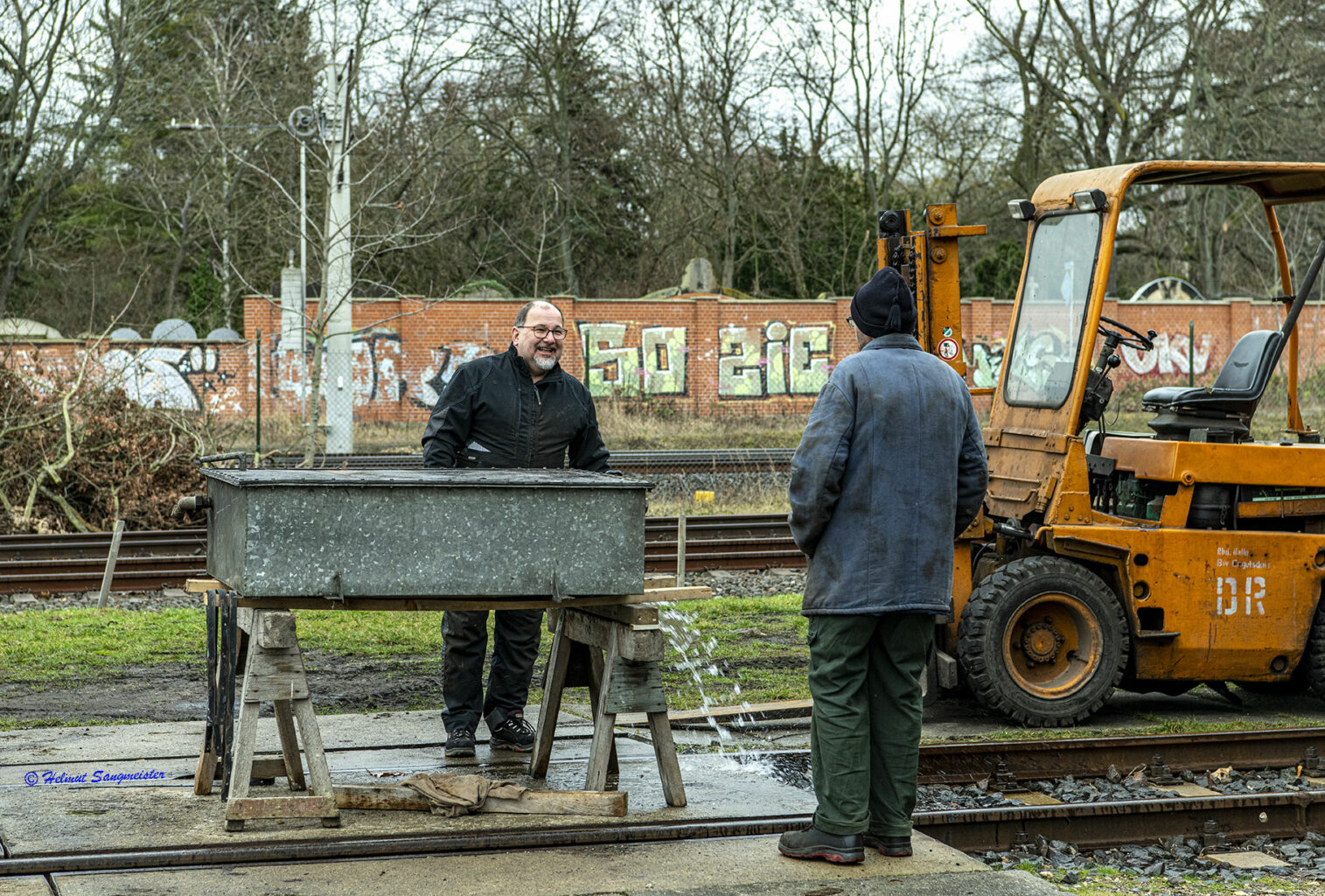 Sie sollten sehen: einen Feuer verzinkten rechteckigen Behälter. Zwei Mitarbeiter beraten über das weitere Vorgehen. An rechten Ende des Bildes steht ein oranger Gabelstabler, mit dem der Behälter transportiert wurde. Der Behälter ist auf Böcken in Arbeitshöhe abgelegt.