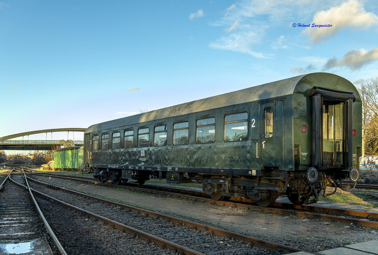 Sie sollten sehen: einen Personenwagen der Gattung Bghw bei blauem Himmel. Der Wagen ist im Originalzustand der Deutschen Reichsbahn der DDR in olivgrün mit grauem Dach. Unter dem fünften Fenster (Mitte) prangt das weiße Logo der DR. Der Wagen verfügt über Drehgestelle mit je zwei Achsen, Bauart Görlitz fünf.