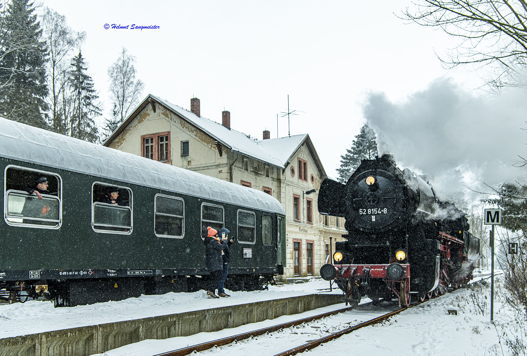 Sie sollten sehen: die Dampflokomotive 52 8154 des Eisenbahnmuseums Leipzig im Bahnhof Holzhau. Es ist winterlich mit etwas Schnee. Links im Bild angeschnitten ein Reisezugwagen der Gattung ABme, der zum Zug gehört. Die Lok umfährt den Zug. Im Hintergrund das Bahnhofsgebäude.