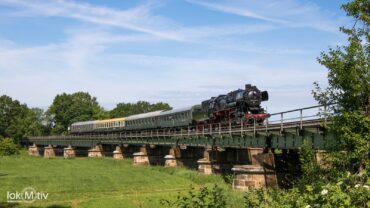 52 8154 überrollt mit dem Museumszug die Auenbrücke in Kössern. Die Brücke ist flach und lang aus Stahl mit Natursteinsäulen.