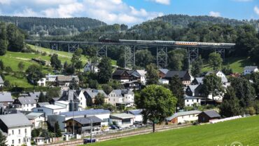 Das Bild zeigt eine Seitenansicht des Markersbacher Viaduktes mit dem Leiziger Museumszug, gezogen von der Dampflokomotive 52 8154. Im Hintergrund das Erzgebirge mit üppiger grüner Vegetation.