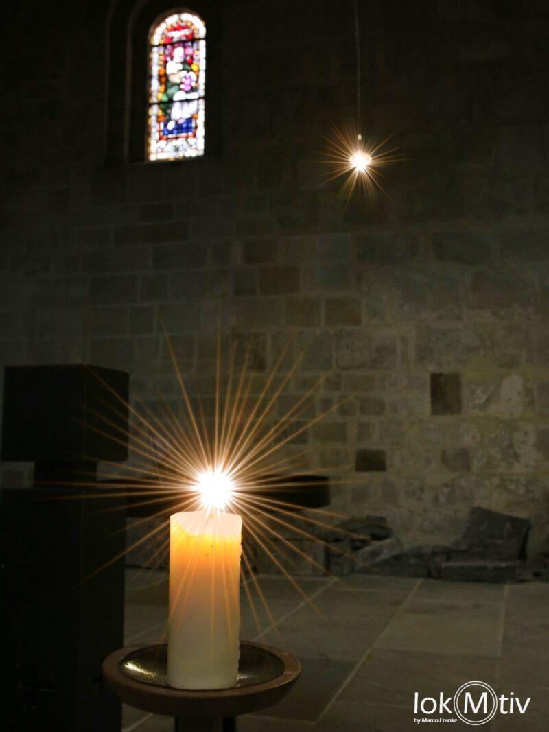 Das Bild zeigt eine leuchtende Kerze und ein kleines kunstvolles Fenster im Hintergrund.