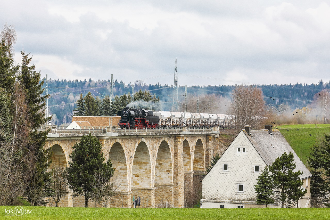 Die vereinseigene Lokomotive 52 8154 auf einem Natursteinviadukt mit großen Rundbögen in Colmnitz. Die Lokomotive befördert einen Güterganzzug. Rechts im Bild ein altes Bauernhaus, giebelseitig zum Betrachter.