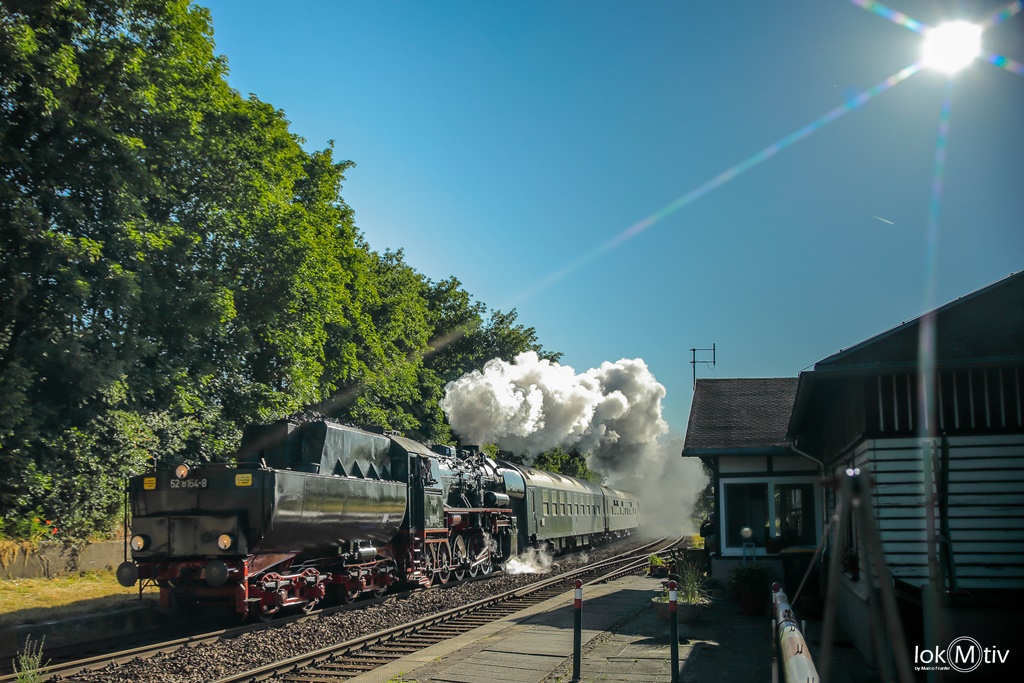 Die Dampflokomotive 52 8154 des Eisenbahnmuseums fährt mit dem Tender voraus im morgentlichen Gegenlicht durch Haynsburg. Eine weiße Dampffahne liegt über dem Zug. Rechts im Bild das hölzerne Bahnhofsgebäude von Haynsburg.