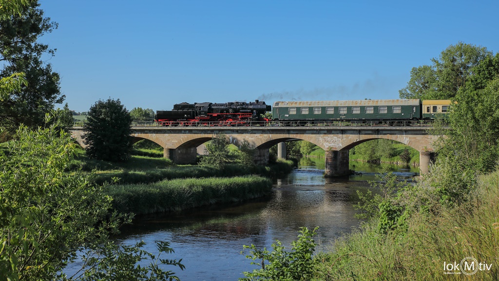 Das Bild zeigt die Dampflokomotive 52 8154, die Tender voraus fahrend, mit einem Teil des Museumszuges auf der Steinbogenbrücke über die ruhig dahinfließende Elster.