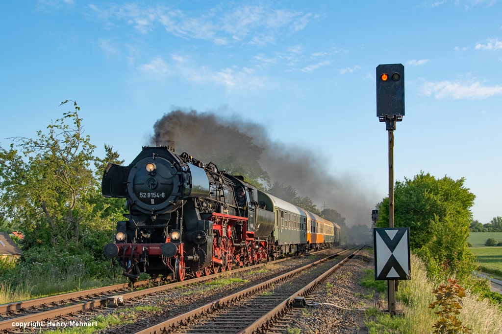 Das Foto zeigt die Dampflokomotive 52 8154 des Eisenbahnmuseums Leipzig im Abendlicht mit dunkler Rauchfahne über dem angehangenem Reisezug. Der Zug passiert ein gelb zeigendes Lichtsignal.