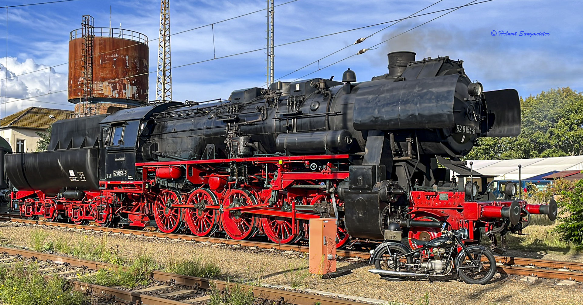Das Bild zeigt die Dampflokomotive 52 8154 und auf Höhe des Vorläufers ein historisches Motorrad. Im Hintergrund der etwas rostige Wasserturm in Falkenberg/Elster.