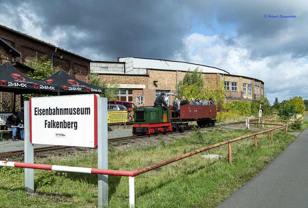 Das Bild zeigt das Eingangsschild des Eisenbahnmuseum Falkenberg. Rechts daneben zu sehen aber räumlich dahinter fährt die Feldbahn mit einer Diesellok und einem Wagen.
