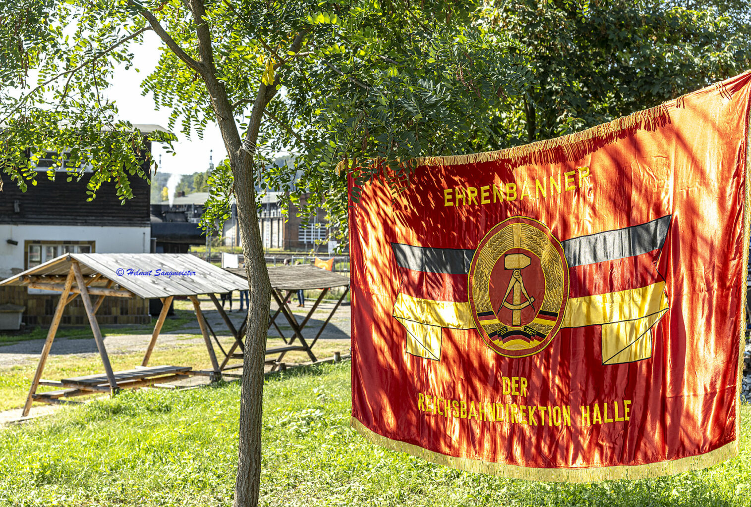 Das Bild zeigt das Ehrenbanner der Reichsbahndirektion Halle. Das Banner ist eine rote Fahne mit dem Symbol der Deutschen Demokratischen Rebublik, also Hammer und Zirkel im Ehrenkranz. Links und rechts sind bandförmig die Farben schwarz, rot, gold zu sehen. Goldener Saum vollendet das Kunstwerk, welches auch die Aufschrift der Rbd Halle trägt.