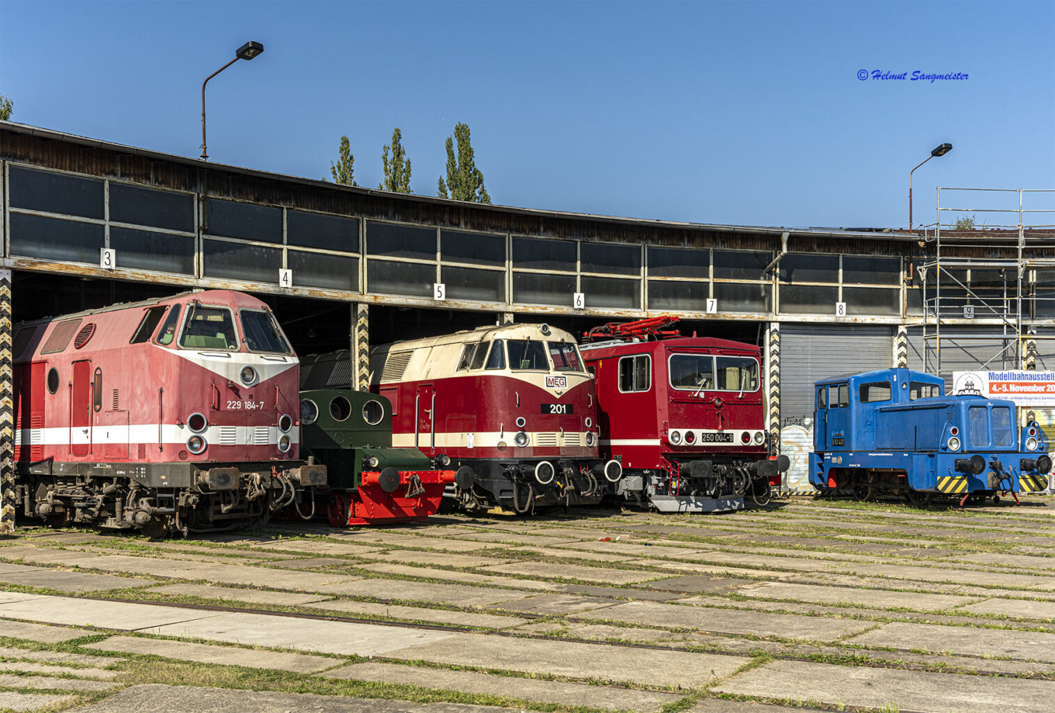 Das Bild zeigt 5 Lokomotiven aus dem Rundhaus der Geraer Eisenbahnwelten schauen. Darunter eine Diesellok der Baureihe 229, auch U-Boot genannt, eine Diesellok der Baureihe 118, auch dicke Babelsbergerin, eine E-Lok der Baureihe 250 der DR, ob ihres kastenförmigen Aussehens auch Energiecontainer oder Kabelcontainer genannt. Ganz rechts eine blaue Kleindiesellok V10.