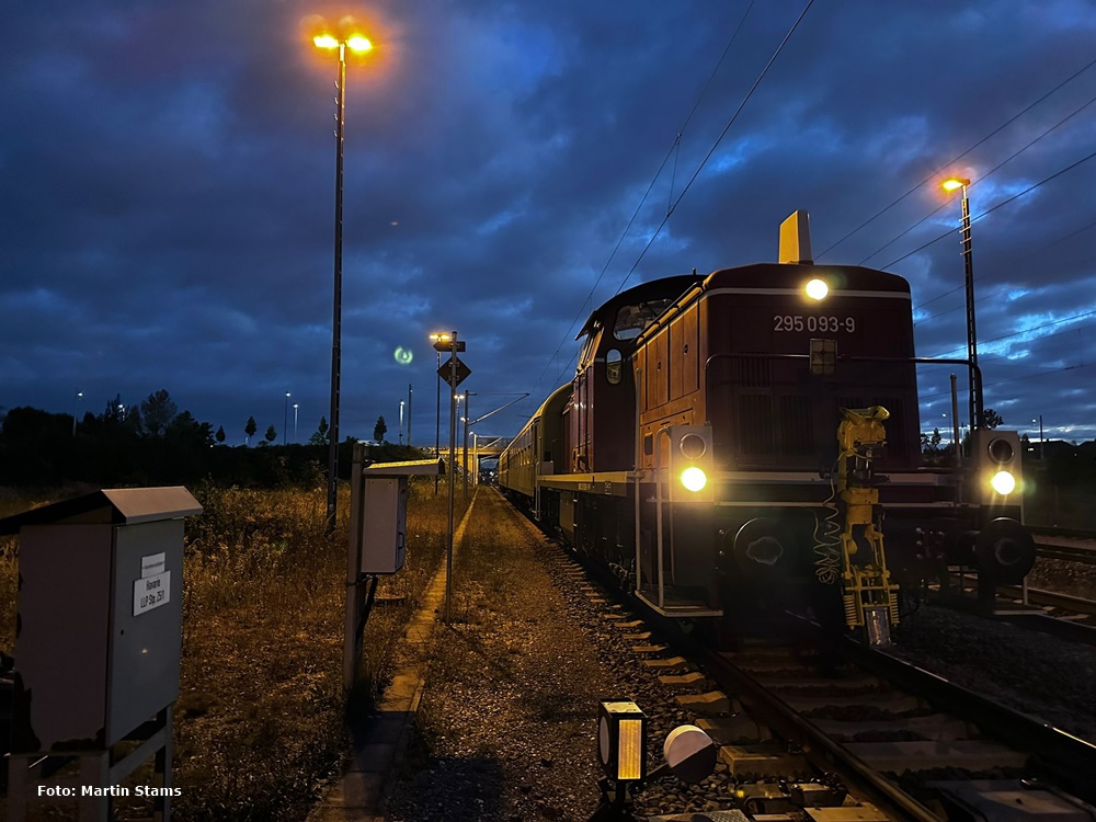 Das Bild zeigt die Diesellok 295 093 in der Morgendämmerung. Beidseitig stehen Laternen und beleuchten die Szenerie.