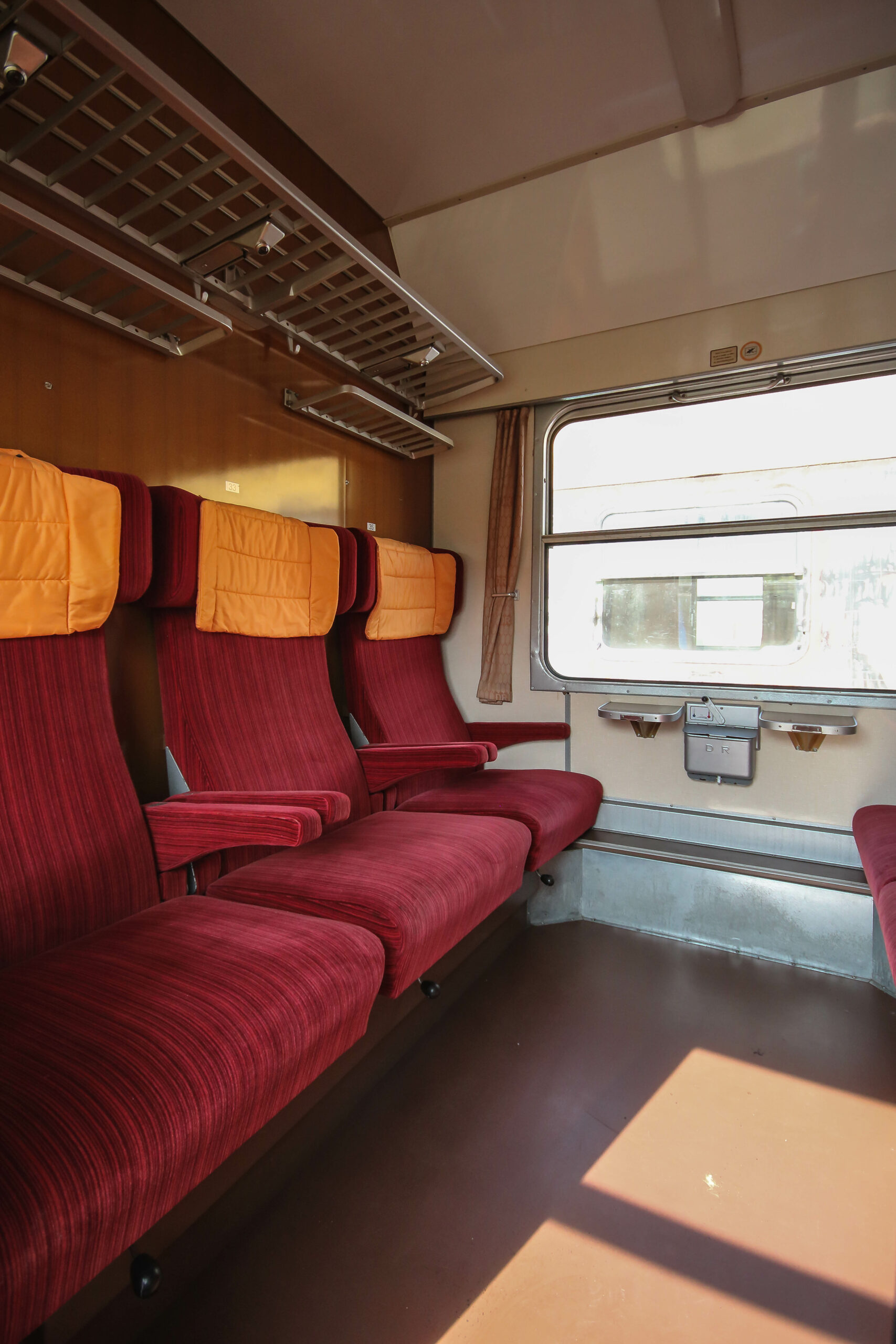 Das Bild zeigt den Blick in ein erste Klasse Abteil. Die drei Sitze einer Reihe verfügen über getrennte Armlehnen, sind mit rotem Stoff bezogen und haben Kopfkissen.