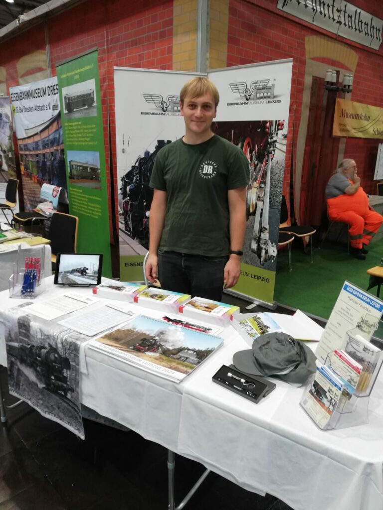 Das Bild zeigt den Messestand des eisenbahnmuseums Leipzig mit Fanartikeltisch, Rollups...