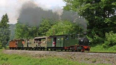 Der sächs. IK Zug mit schwarzer Rauchfahne auf gerader Strecke. Der Zug besteht aus verschiedenen Wagen, darunter einem offenen Wagen mit Planendach.