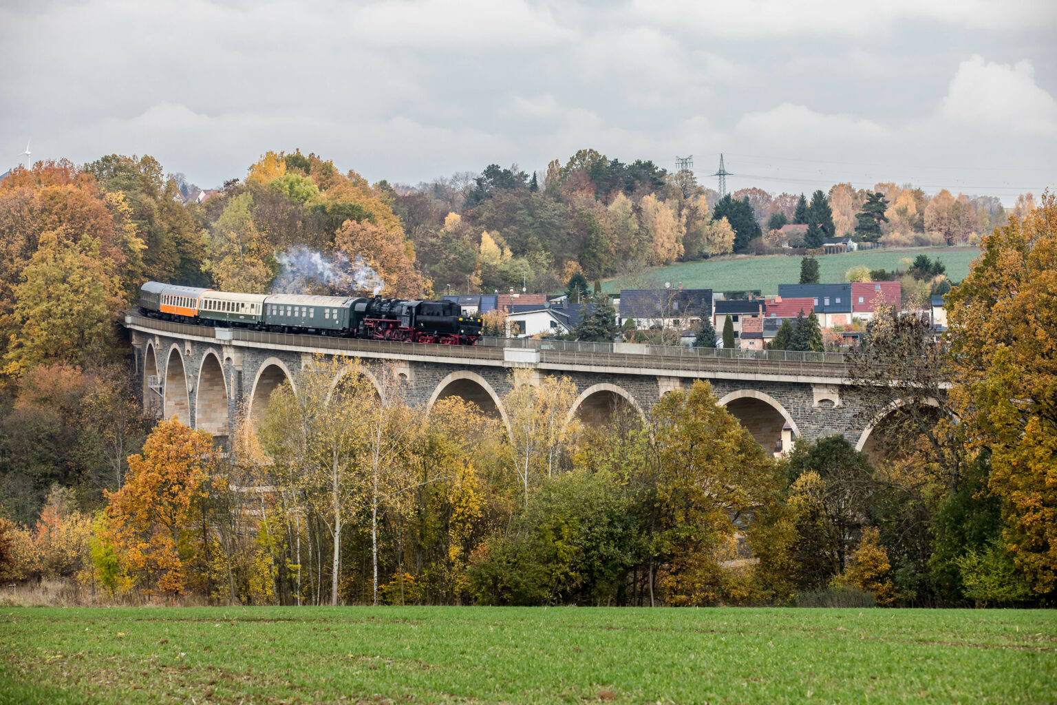 Das Bild zeigt den Zug des Eisenbahnmuseums Leipzig auf dem Bahrebachmühlenviadukt bei Chemnitz. Die Dampflokomotive hängt mit dem Tender voran am Zug. Der Natursteinviadukt ist von herbstlichen Bäumen umgeben.