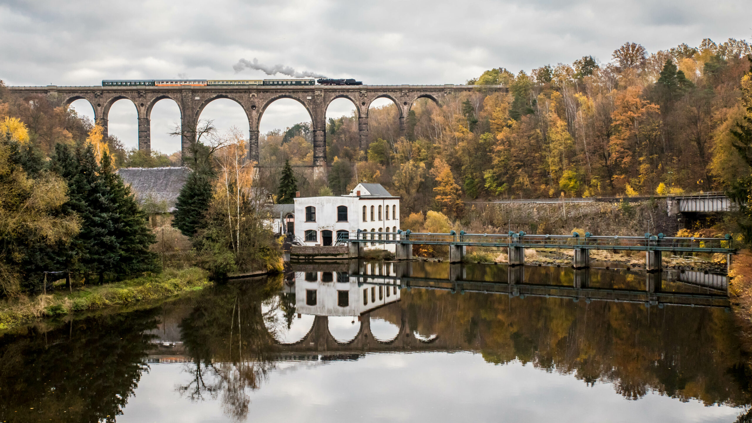 Zu sehen ist der Museumszug des Eisenbahnmuseums Leipzig auf dem Naatursteinviadukt in Göhren. Der Viadukt spiegelt sich inkl. des Zuges im aufgestauten Wasser eines Wehrs vor dem Viadukt.