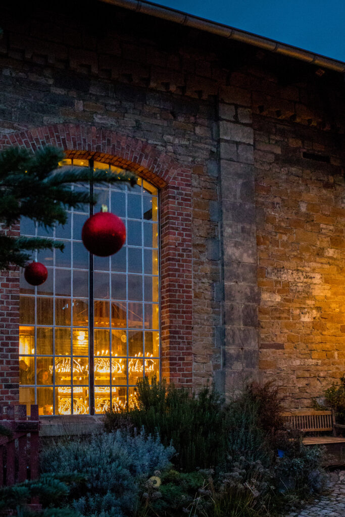 Das Bild zeigt einen mit roten Kugeln geschmückten Ast des Weihnachtsbaums vor einem festlich beleuchteten Fenster des Lokschuppens.