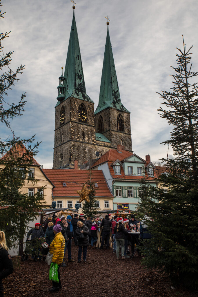 Eingerahmt von zwei Tannen geht es auf einen der vielen Plätze, die als Weihnachtsmarkt genutzt sind. Im Hintergrund thronen zwei Kirchtürme.