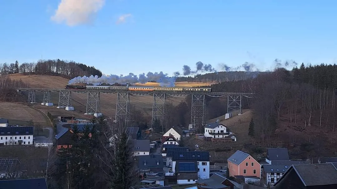 Unser Zug mit langer Dampffahne im Licht auf dem Markersbacher Viadukt. Im Vordergrund die Gemeinde Markersbach eingebettet in die typisch erzgebirgische Hügellandschaft.