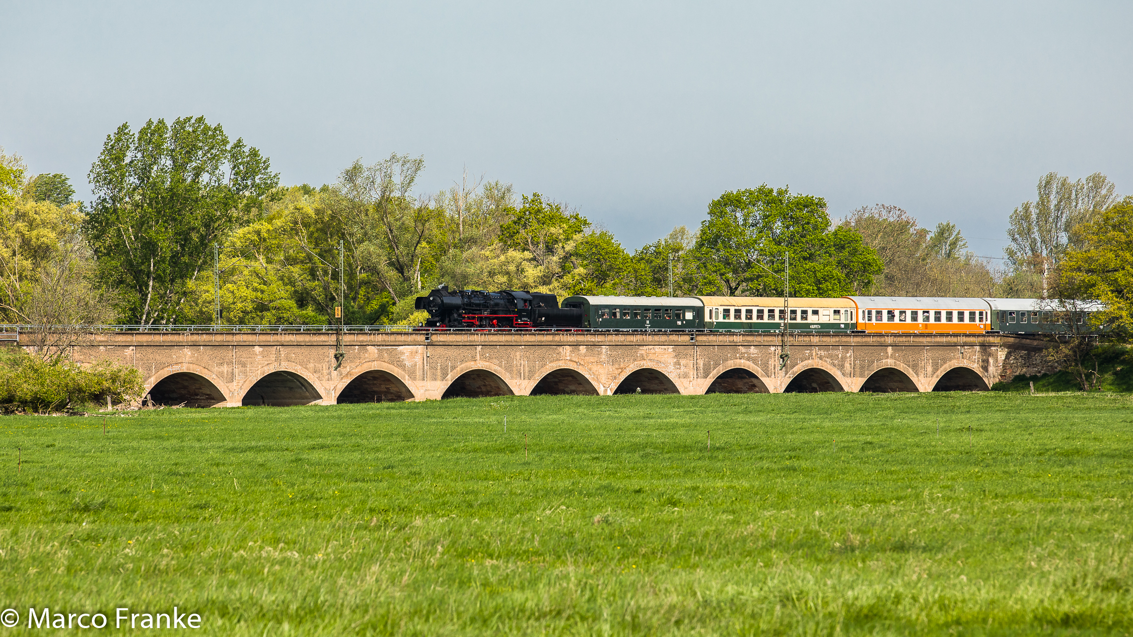Der Zug auf einer steinernen Bogenbrücke im Grün der Landschaft. Kaum zu erahnen, dass sich wenige Hundert Meter weiter Schornsteine gen Himmel strecken.