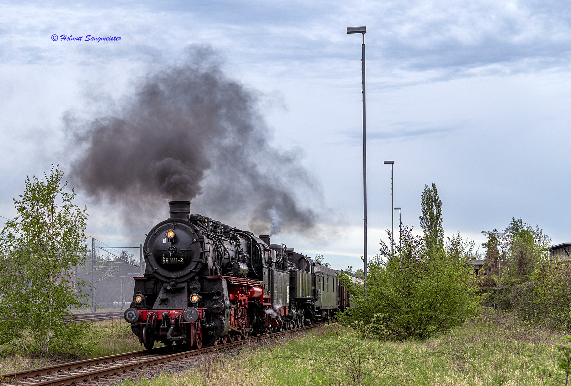 Dampflok 77.250 als Zuglok mit Vorspann durch 58.1111 verlassen mit dunkler Qualmfahne das Eisenbahnmuseum Leipzig. Das Bild zeigt den Zug zwischen bereits grünenden Streuchern und kleinen Birken.