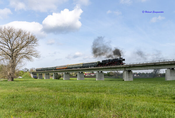 Der komplette Zug mit kleiner schwarzer Qualmwolke auf der Neubaubrücke über die Aue in Kriechau. Die Brücke ist aus Beton, schnurgerade und der Zug hebt sich toll gegen den blauen Himmel ab.