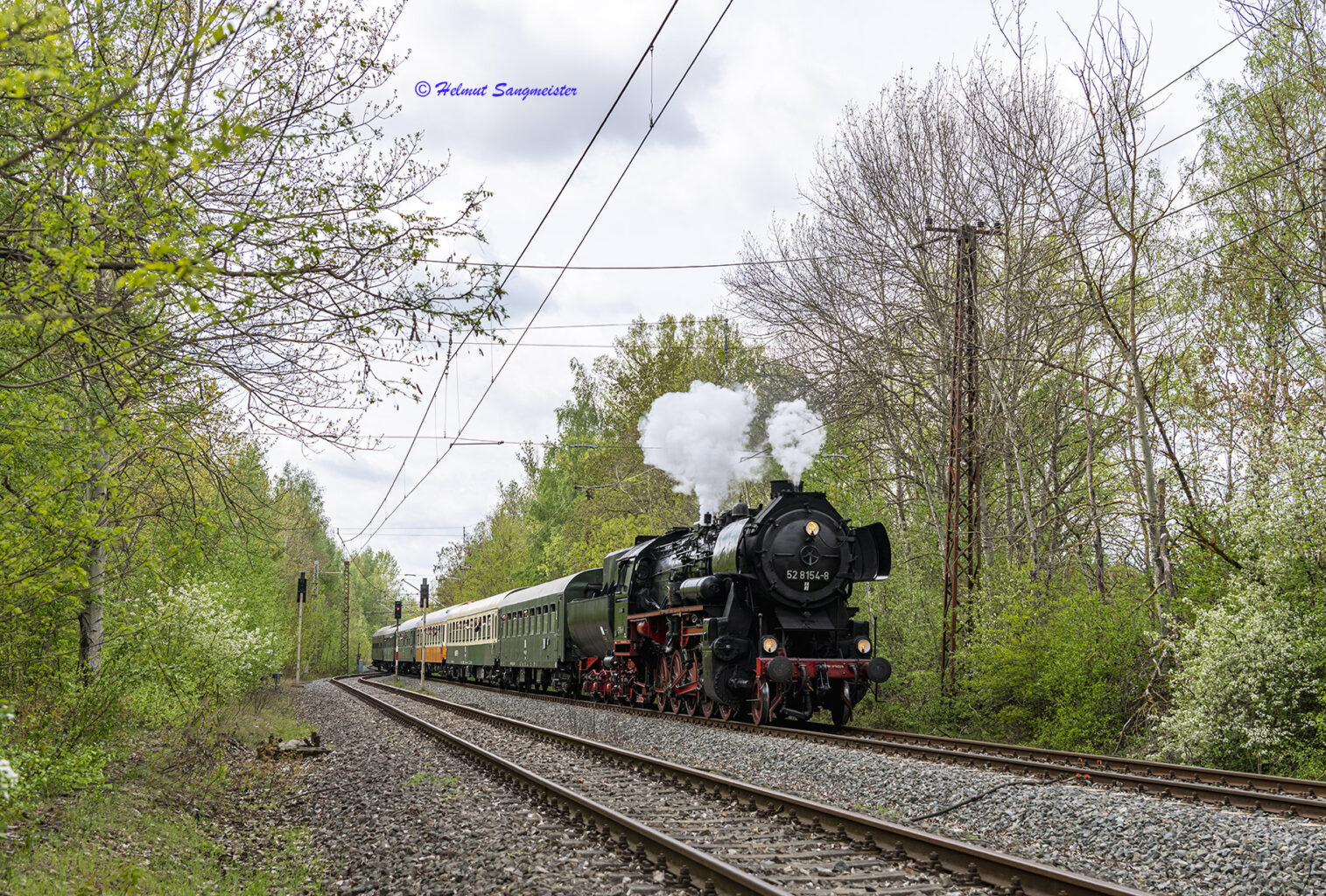 Dampflok 52 8154 des Eisenbahnmuseums Leipzig mit dem voll besetzten Reisezug auf einem der zweigleisigen Abschnitte vor dem Bahnhof Profen. Über der Dampfpfeife weißer Dampf, ein Zeichen, dass es laut wird. Neben dem zug die typischen Lichtsignale der MIBRAG Infrastruktur.